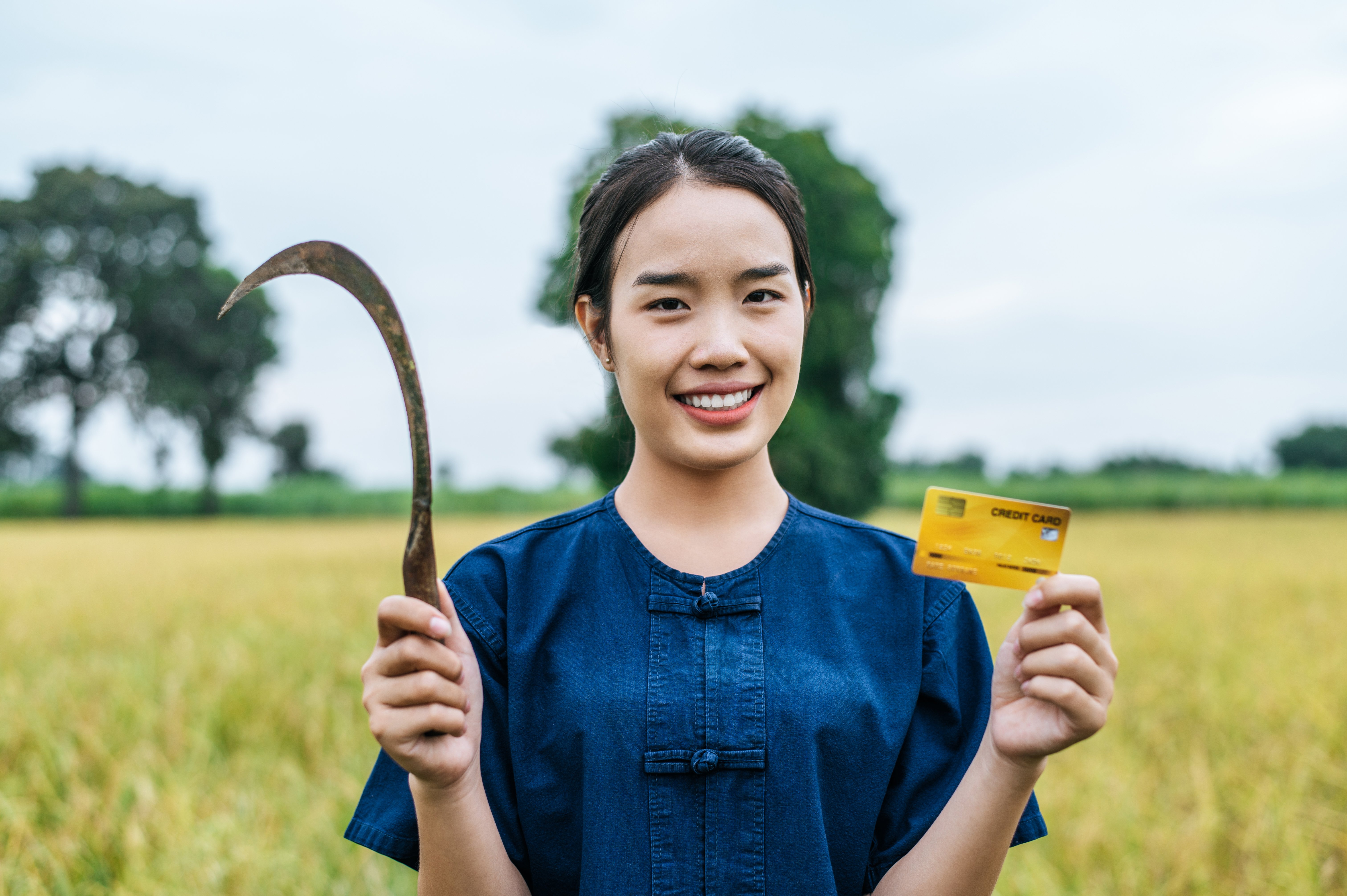 Mulher asiática segurando uma foice em uma mão e um cartão de crédito em outra mão, ela está sorrindo e usando uma roupa azul, ao fundo, uma plantação de soja e uma árvore.
